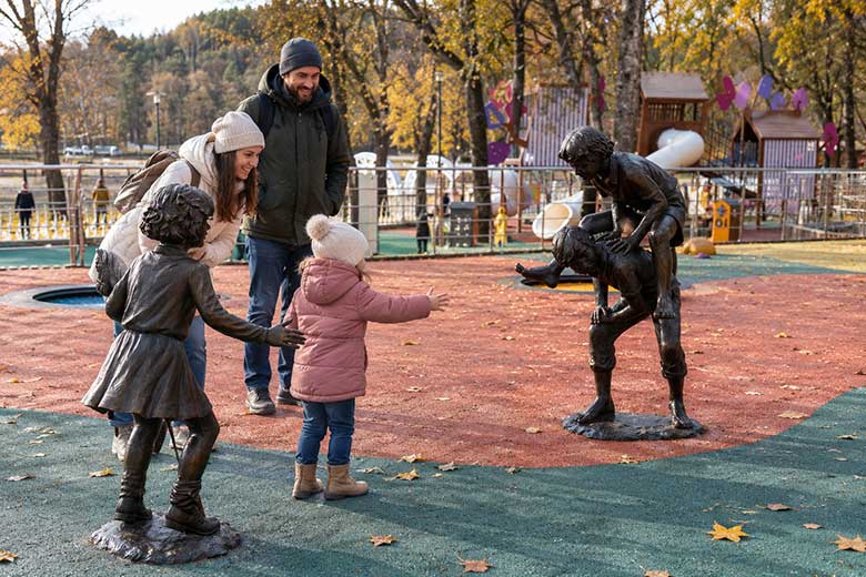 Семья с маленьким ребёнком гуляет по осенней площадке среди бронзовых скульптур детей.