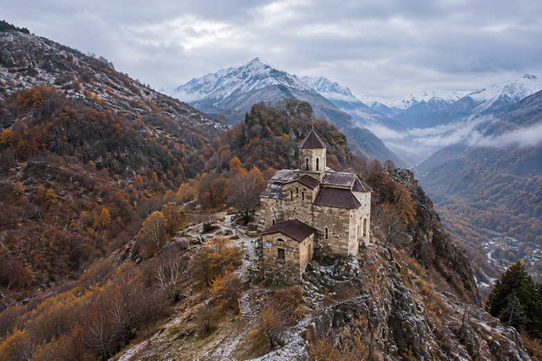 Старинная каменная церковь на горном хребте, осенний лес с первым снегом и заснеженные вершины.