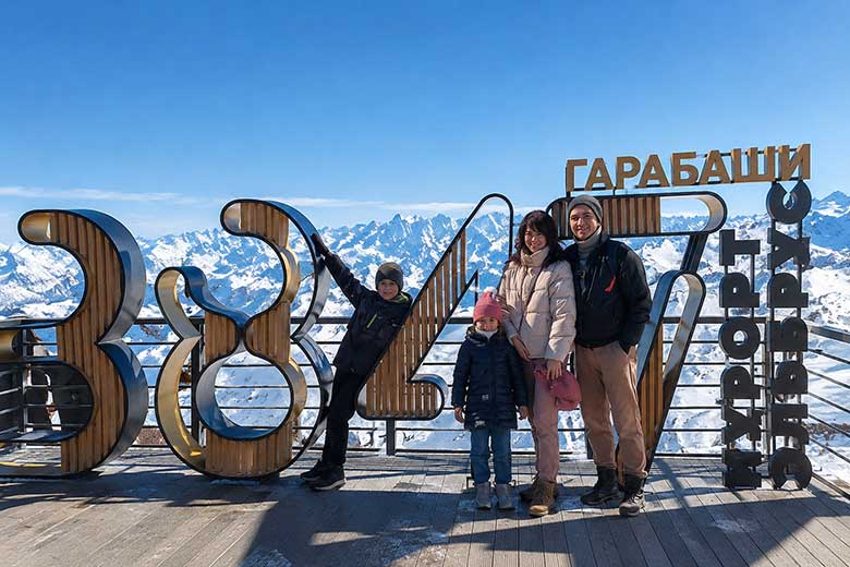 Семья с детьми фотографируется на смотровой площадке Гарабаши на высоте 3847 метров на фоне заснеженных гор Кавказа.